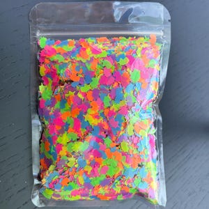 May include: A clear plastic resealable pouch filled with a large quantity of small, flat, unicorn-shaped confetti pieces in vibrant neon colors including pink, orange, yellow, green, and blue, creating a dense and colorful multi-colored display.