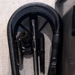 May include: A black vacuum cleaner with various attachments, including a brush and a crevice tool, is neatly stored in a compartment. The vacuum's hose is coiled, and the metal wand is visible. The unit is designed for efficient cleaning.