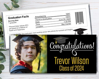 May include: A graduation chocolate bar with a photo of a graduate in a cap and gown. The bar is black with gold lettering that says "Congratulations! Trevor Wilson Class of 2024".