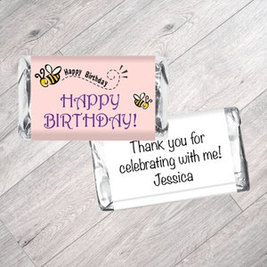 May include: Two small chocolate bar wrappers with a pink background and bee illustrations. The top wrapper says "Happy Birthday!" and the bottom wrapper says "Thank you for celebrating with me! Jessica".