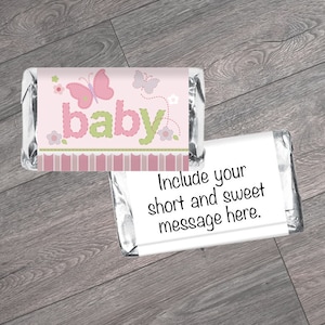 May include: Pink and green baby shower favor chocolate bar wrapper with a butterfly design and the word "baby" in a stitched font. The wrapper has a space for a short message.