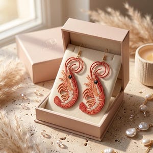 May include: A pair of drop earrings featuring embroidered shrimp in shades of orange and red. Each shrimp has a black eye and is suspended from a gold-toned metal hook. The earrings are displayed inside an open, light pink rectangular gift box.