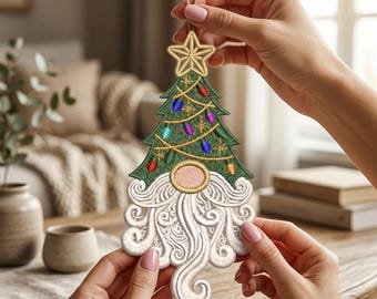 Pode incluir: Um enfeite de árvore de Natal suspenso com uma árvore verde bordada, uma estrela dourada no topo, luzes coloridas e um gnomo com uma grande barba branca em espiral e um nariz redondo cor de pêssego, suspenso por um cordão de juta.