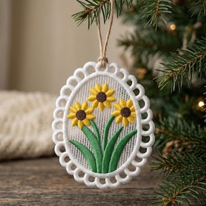 Pode incluir: Enfeite de Natal oval branco com borda ondulada, apresentando três girassóis bordados. Os girassóis têm pétalas amarelas e centros marrons, com folhas e caules verdes. Ele pende de uma corda de barbante natural.