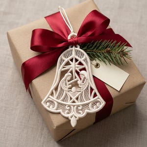 May include: A cream-colored lace bell ornament featuring a nativity scene with a star at the top. The ornament is attached to a brown gift box wrapped with a deep red satin ribbon, a small sprig of pine needles, and a blank rectangular gift tag.