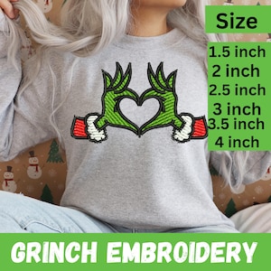 Könnte beinhalten: Ein hellgraues Sweatshirt mit einem grünen, gestickten Herz, das aus Grinch-Händen geformt ist. Die Hände haben weiße Manschetten und rote Ärmel. Der Text "GRINCH EMBROIDERY" steht unten. Größen von 3,8 cm bis 10,2 cm.