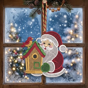 May include: A hanging Christmas ornament featuring an embroidered Santa Claus with a white beard and red hat, positioned next to a yellow birdhouse with a green roof and a small red bird perched on top, suspended by a red and gold twisted cord.