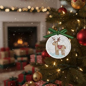 Könnte beinhalten: Ein runder Weihnachtsschmuck mit einem gestickten Rentier-Design. Der Schmuck, mit einem grünen Band, hängt an einem Weihnachtsbaum. Das Rentier ist braun mit einer rot-weiß gemusterten Decke. Der Hintergrund umfasst einen Kamin und verpackte Geschenke.
