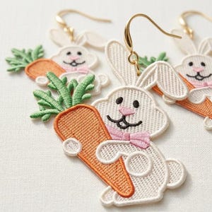 Könnte beinhalten: Bestickte Hasenohrringe mit Karotte. Die weißen Hasen haben schwarze Augen, rosa Nasen und rosa Schleifen. Die Karotten sind orange mit grünen Blättern. Die Ohrringe haben goldfarbene Haken.