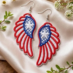 May include: A pair of patriotic wing-shaped earrings. Each earring features a red and white striped wing design with a blue section containing a white star. The earrings have silver-colored hooks and are set against a satin background.