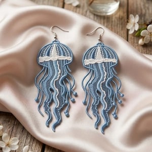 May include: A pair of dangling earrings featuring embroidered jellyfish designs in shades of light blue and white. Each earring has a silver-toned metal hook and a detailed thread-stitched body with long, flowing tentacles that end in small knots.