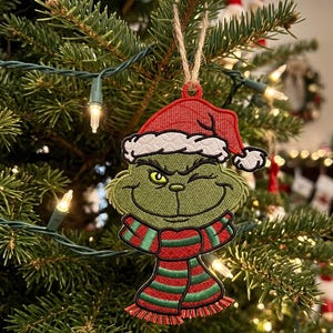 Puede incluir: Adorno bordado para árbol de Navidad que representa al Grinch, con gorro de Papá Noel rojo y bufanda a rayas. El personaje verde guiña un ojo. El adorno cuelga de una cuerda en un árbol de Navidad decorado con luces.