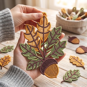 Può includere: Un ornamento ricamato raffigurante una ghianda centrale con cupola marrone scuro e frutto giallo dorato, circondato da un gruppo di foglie di quercia dettagliate in tonalità giallo oro e verde salvia con venature e bordi marrone scuro.