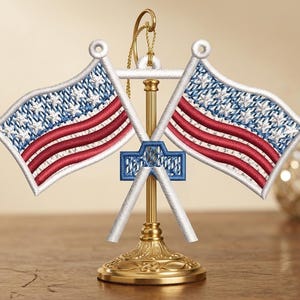May include: Embroidered American flag ornament featuring two flags with red and white stripes and blue star fields. The flags are crossed and mounted on a gold-colored base, with a gold hanging loop.