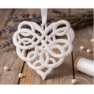 Op de afbeelding: Een wit, geborduurd hartvormig ornament met een ingewikkeld, verweven ontwerp. Het ornament hangt aan een wit lint. Verspreide parels en een spoel wit garen liggen op een houten oppervlak, met gedroogde lavendel op de achtergrond.