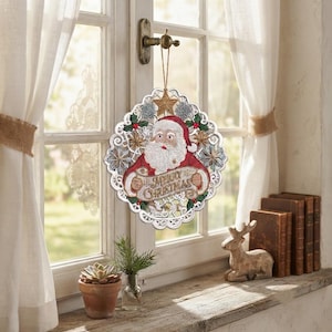 Pode incluir: Um enfeite de Natal circular suspenso com um Pai Natal bordado em fato vermelho segurando uma faixa com o texto Merry Christmas. O design &eacute; rodeado por uma borda branca tipo renda com detalhes de estrelas e flocos de neve em dourado e prateado.