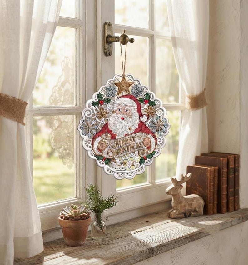 Pode incluir: Um enfeite de Natal circular suspenso com um Pai Natal bordado em fato vermelho segurando uma faixa com o texto Merry Christmas. O design &eacute; rodeado por uma borda branca tipo renda com detalhes de estrelas e flocos de neve em dourado e prateado.