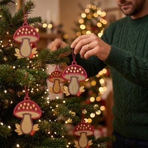Pode incluir: Enfeites de árvore de Natal em forma de cogumelo com um chapéu vermelho escuro com pontos amarelo-claros, um caule bege e folhas douradas na base. Cada enfeite pendura-se num fio vermelho fino e apresenta um design texturado bordado.