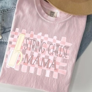 May include: A light pink t-shirt with the words "STRING CHEESE MAMA" in a playful font, set against a pink and white checkered background. A cartoon string cheese graphic is on the left. The shirt is folded on a denim surface.
