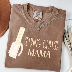 May include: Brown t-shirt with the words "STRING CHEESE MAMA" in a light beige font. The shirt features a graphic of a string cheese stick with a white wrapper. A silver necklace with a small pendant is also visible.