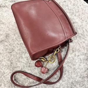 Pode incluir: Uma bolsa de ombro de couro rosa empoeirado com acabamento liso, apresentando um fecho de zíper superior e uma alça de couro fina combinando. Um chaveiro de metal dourado com dois pingentes de cereja brilhantes, um vermelho escuro e um rosa claro.