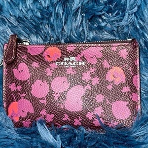 May include: A Coach key pouch with a floral pattern in pink and orange on a dark brown background. The pouch has a zipper closure, a silver-tone key ring, and the Coach logo in silver.