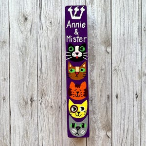 A vertical wooden sign with a purple background and a crown at the top, featuring four cat faces with different expressions.