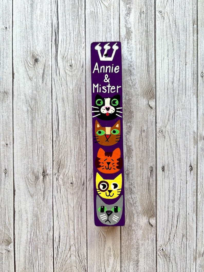 A vertical wooden sign with a purple background and a crown at the top, featuring four cat faces with different expressions.