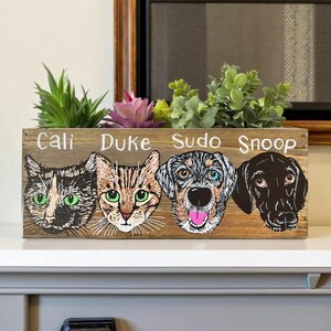 A wooden planter box with four illustrated animal faces, each labeled with a different pet's name, placed on a shelf with a potted plant in the background.