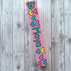 A pink bookmark with a crown and hearts on it, placed on a wooden surface.