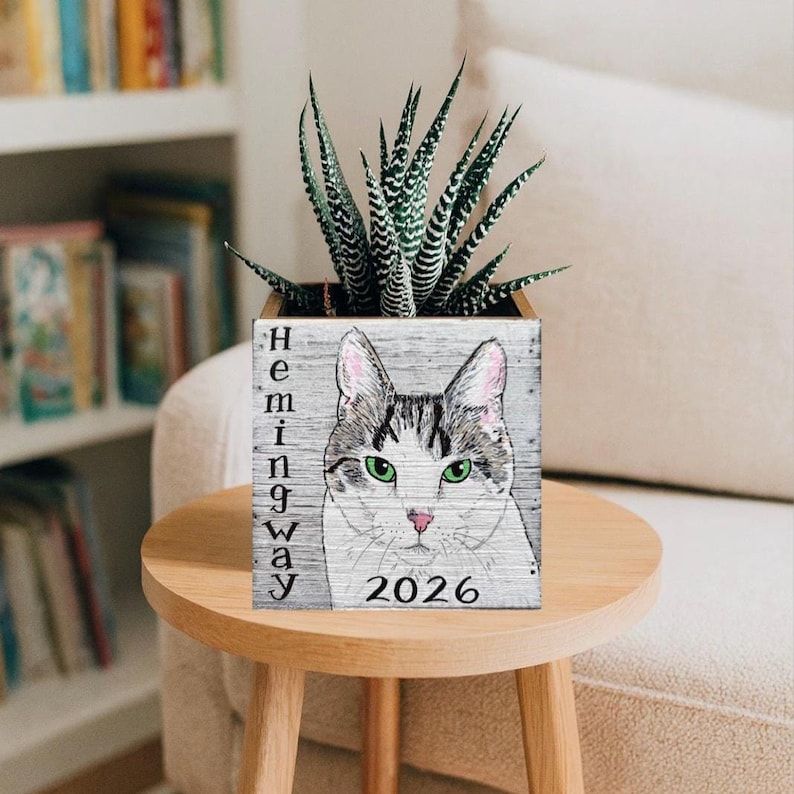 May include: A wooden planter with a cat portrait and the name "Hemingway" vertically on the side. The cat has green eyes and a pink nose. The year "2026" is below the cat. The planter holds a succulent plant.