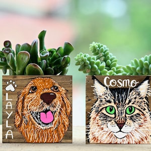 May include: Two wooden planters with hand-painted pet portraits. One features a golden retriever with the name "Layla" and a paw print. The other depicts a cat with green eyes, named "Cosmo". Both planters hold green succulents.