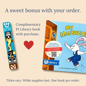 The image is an advertisement for a library service that offers a sweet bonus with an order. It features a book titled "My Mezuzah" and a bookmark with a cartoon rabbit. The text on the image provides information about the library service, including that it is a complimentary PJ Library book with purchase, and that the books are available for one book per order. The image also mentions that the titles vary, and that while supplies last, one book per order is the standard.