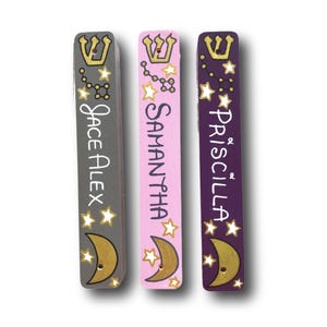 May include: Three rectangular mezuzah cases in gray, light pink, and dark purple. Each features a gold Hebrew letter Shin at the top, a constellation, white stars, and a gold crescent moon at the bottom. Names Jace Alex, Samantha, and Priscilla are printed.