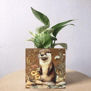 May include: A square wooden planter with a painted otter illustration, surrounded by flowers and greenery. The planter holds a green and white leafy plant. The background is a light wood surface.