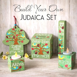 May include: A set of light green wooden Judaica items featuring hand-painted trees, pomegranates, and blue birds. The set includes a hamsa wall hanging, a square tzedakah box, a tall mezuzah case, and two square candle holders with tea lights.