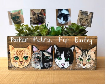 May include: A wooden planter box with four painted cat faces and corresponding photos above. The names "Parker", "Petra", "Pip", and "Bailey" are written above each cat. The planter is filled with green succulents.