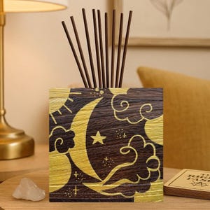 May include: A decorative wooden aromatherapy diffuser with a celestial design featuring a crescent moon, stars, and clouds in gold against a dark brown background. The diffuser holds several brown reed sticks, adding a touch of natural fragrance to any space.