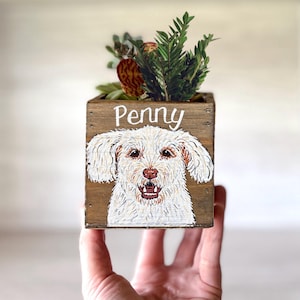 May include: A wooden planter box featuring a hand-painted portrait of a white dog. The dog's name, "Penny," is written above the image in white script. The box is filled with a variety of green and red succulents.