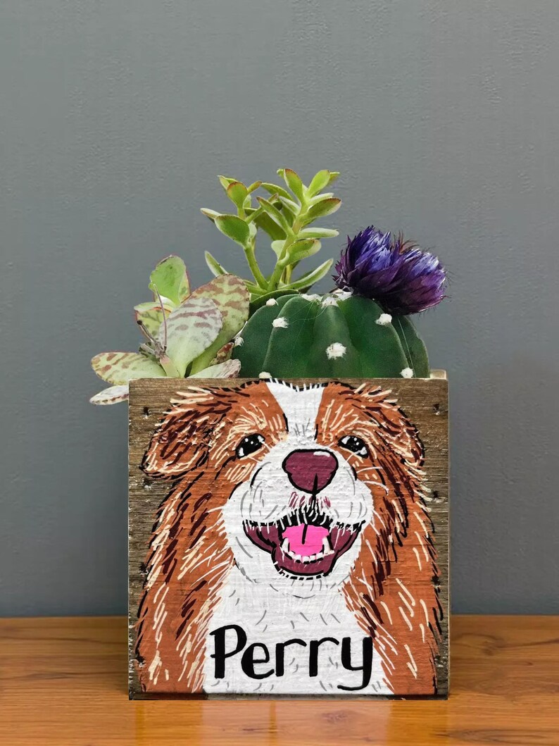 A wooden planter box with a painted illustration of a dog's face, featuring the name "Perry" below it. The planter box is filled with various plants, including a cactus and a purple flower.