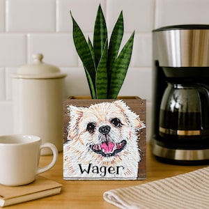 May include: A wooden planter box with a painted portrait of a smiling dog named "Wager." The planter holds a green snake plant. A white coffee cup and a coffee maker are in the background.
