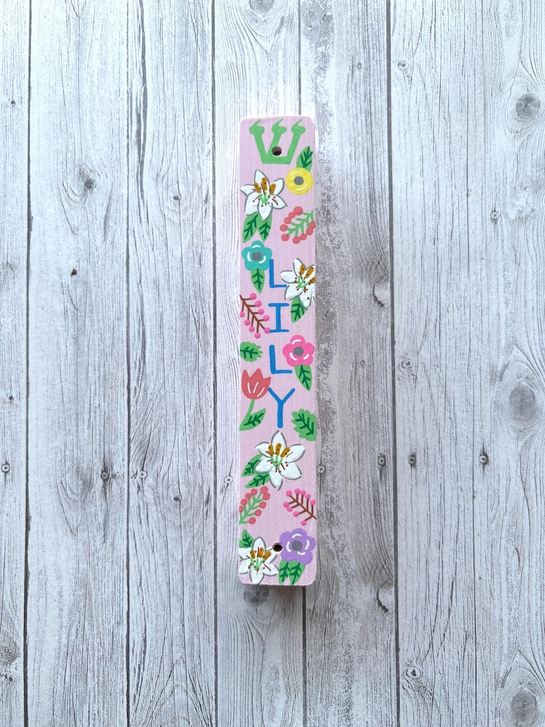 A colorful, hand-painted wooden stick or sign with floral designs and the name "Lily" written on it, placed on a wooden background.