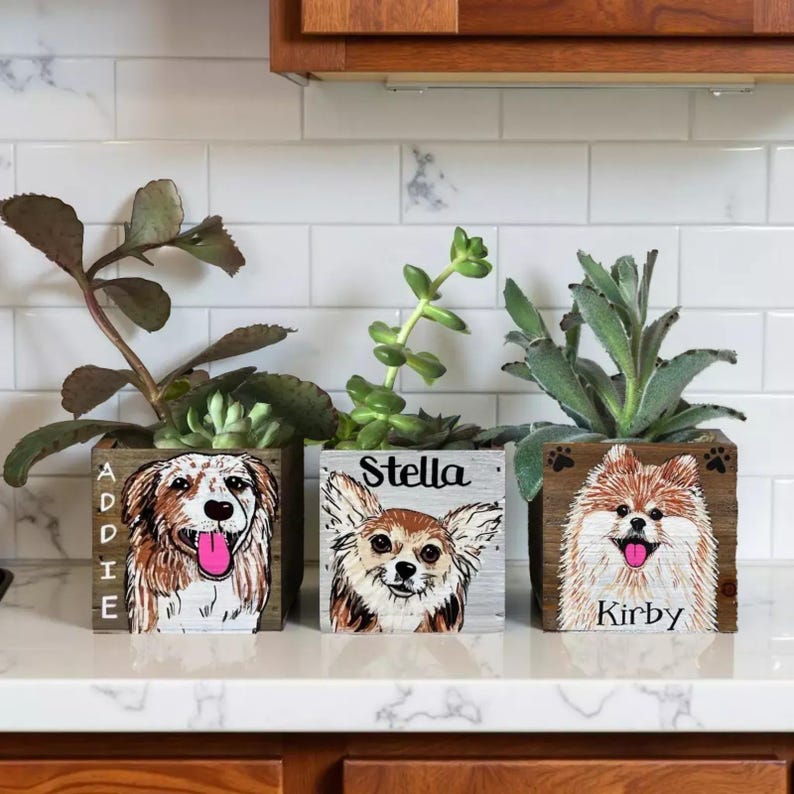 Three wooden planters with painted images of dogs, each with a different name written on them, placed on a white countertop against a tiled wall.