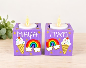 May include: Two purple wooden cube candle holders, each topped with a white tea light. The left cube features the name Maya, an ice cream cone, and a rainbow. The right cube displays Hebrew text, a rainbow, and an ice cream cone with gold stars.