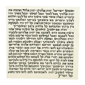 May include: A square parchment featuring black Hebrew text printed in a traditional serif font. The text consists of the Shema Yisrael and V'ahavta prayers, arranged in multiple lines across the surface of the off-white material.