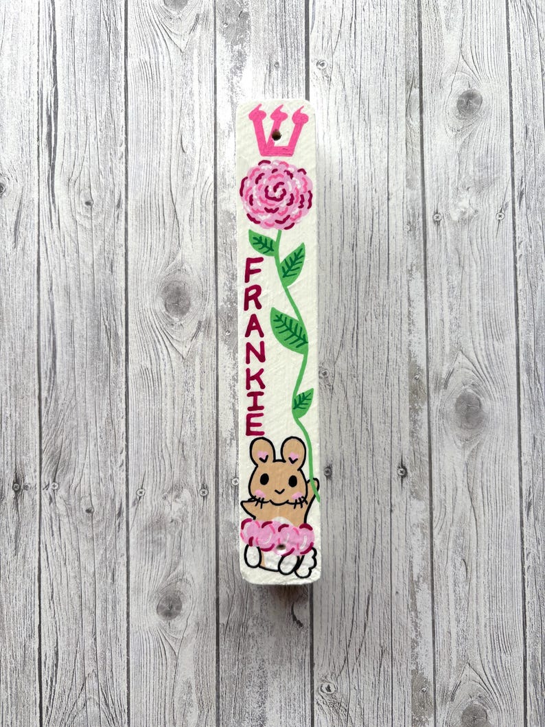 A white wooden board with a pink flower and a cartoon mouse on it, placed against a wooden background.