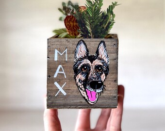 May include: A wooden planter box with a hand-painted German Shepherd dog portrait and the name "MAX" in white letters. The box is filled with a variety of green and red succulents and greenery. The planter is held in a hand.
