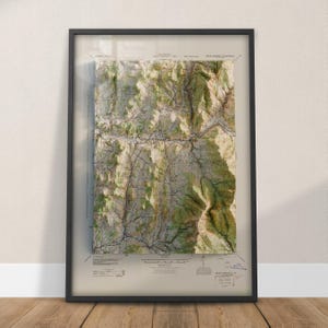 May include: Framed topographic map with a black frame. The map features detailed terrain with shades of green, brown, and beige, depicting mountainous regions. The map includes contour lines, roads, and text labels, offering a detailed geographical representation.