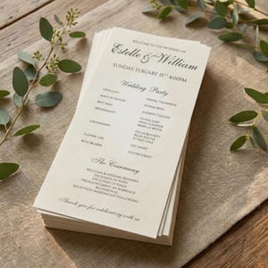 May include: A stack of ivory wedding programs with black text. The programs list the wedding party, ceremony details, and a thank you message. Greenery and a neutral linen cloth are in the background.