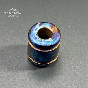 May include: A cylindrical bead with a dark blue and gold iridescent finish. The bead has a hole through the center and gold bands around the top and bottom. The text "GRAIN & METAL CRAFTS" is in the upper left corner.